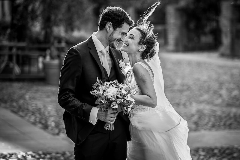 In questa foto in bianco e nero di Nicola Tanzella due sposi su un prato si abbracciano e sorridono. Lo sposo tiene un bouquet 